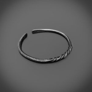 Op de afbeelding: Een zilverkleurige manchetarmband met een gedraaid touwontwerp aan één kant. De open armband heeft een gehamerde textuur en een eenvoudige, elegante stijl. De sieraden worden op een grijze achtergrond getoond.