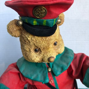 May include: A plush teddy bear dressed as a drummer, wearing a red and green uniform with a black hat and a gold button. The bear is holding a drum and has bells on its uniform.