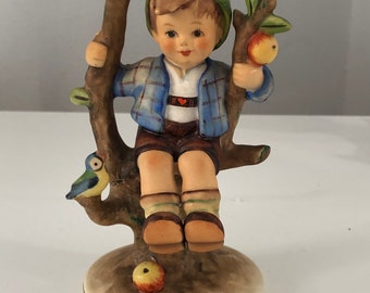 May include: A porcelain figurine of a boy sitting in an apple tree. He is wearing a blue and white plaid shirt, brown pants, and a green hat. There is a bluebird perched on a branch of the tree.