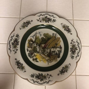 May include: A white ceramic plate with a green border and a floral design. The center of the plate features a painting of a quaint village with houses and a church.
