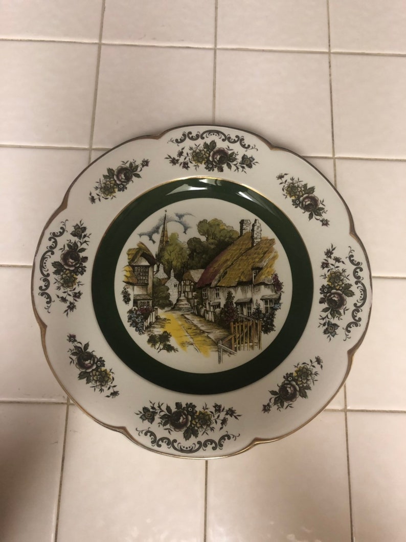 May include: A white ceramic plate with a green border and a floral design. The center of the plate features a painting of a quaint village with houses and a church.