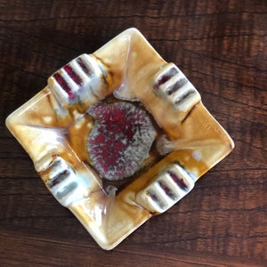 May include: Square ceramic ashtray with a red, white, and brown glaze. The ashtray has four indentations for cigarettes.