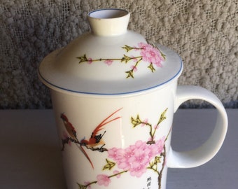 Puede incluir: Taza de té de porcelana blanca con tapa, con un diseño floral con flores rosas y dos pájaros rojos posados en una rama. La taza tiene un borde azul y una asa. La tapa tiene un diseño floral similar. El texto en la taza dice "春風", que se traduce como "Viento de primavera".