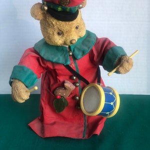 May include: A plush teddy bear dressed as a drummer, wearing a red hat with gold trim, a green and red jacket, and holding a drum with blue and yellow stripes. The bear has a small deer figurine in its pocket.
