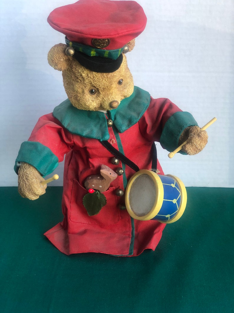 May include: A plush teddy bear dressed as a drummer, wearing a red hat with gold trim, a green and red jacket, and holding a drum with blue and yellow stripes. The bear has a small deer figurine in its pocket.