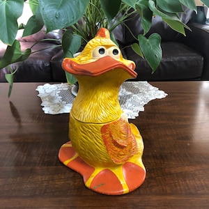 Könnte beinhalten: Eine gelbe Keramik-Entenfigur mit orangefarbenen Akzenten und großen Augen. Die Ente sitzt auf einer Holzoberfläche.