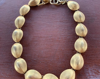 May include: A gold-toned necklace featuring large, textured oval beads. The necklace has a toggle clasp and a series of connected oval-shaped beads. The jewelry is displayed on a brown surface.