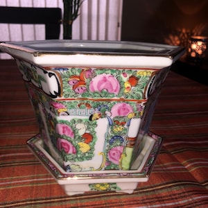 May include: A square, white porcelain planter with a matching saucer. The planter features a colorful floral design with pink roses, green leaves, and gold trim. The design is repeated around the planter's sides.