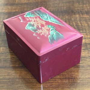 May include: A small, rectangular wooden box with a red lacquered finish. The lid features a painted scene of a mountain range with a pagoda in the background. The scene is painted in shades of green, gold, and white.