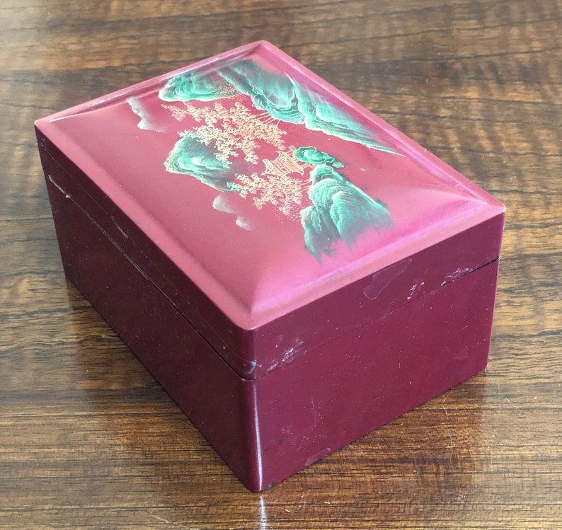 May include: A small, rectangular wooden box with a red lacquered finish. The lid features a painted scene of a mountain range with a pagoda in the background. The scene is painted in shades of green, gold, and white.