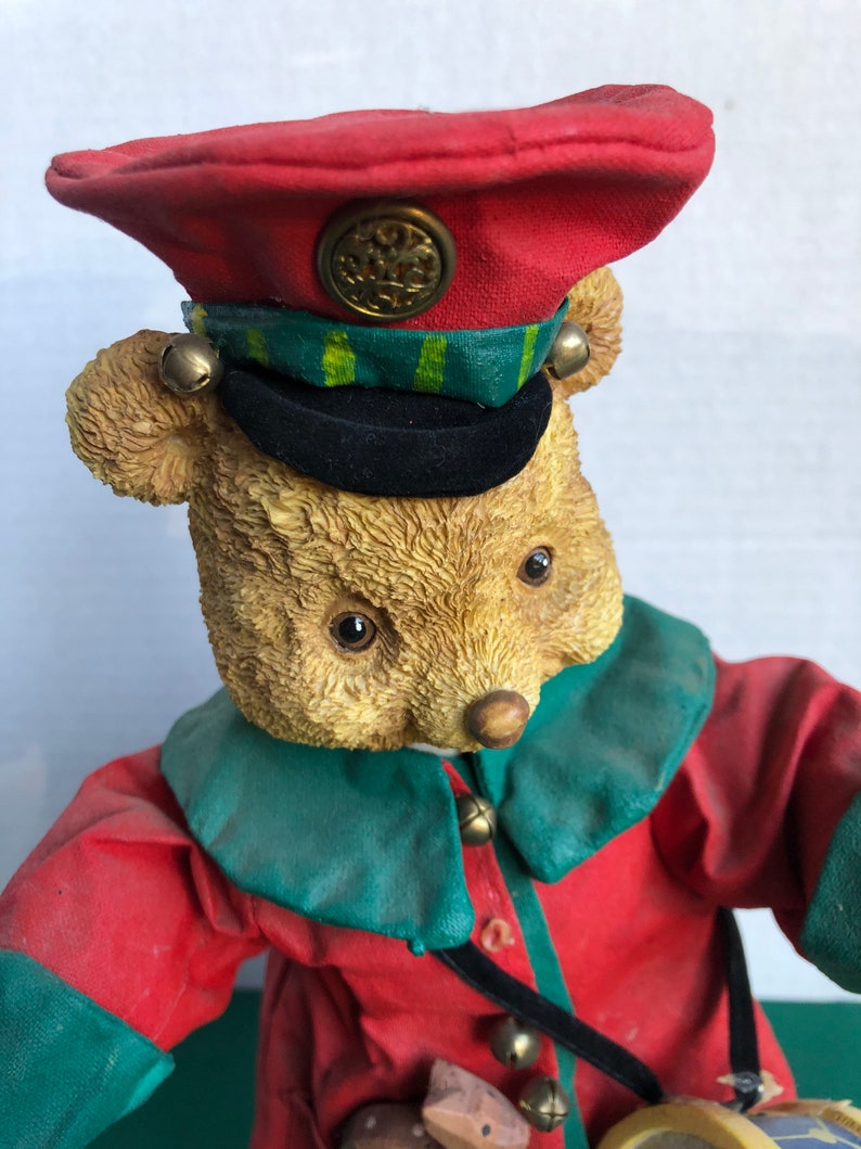 May include: A plush teddy bear dressed as a drummer, wearing a red and green uniform with a black hat and a gold button. The bear is holding a drum and has bells on its uniform.
