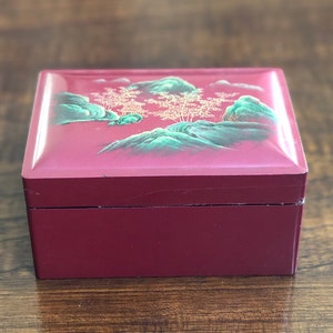 May include: A small, square, red wooden box with a painted scene of green mountains and trees on the lid. The box has a gold trim around the edges.