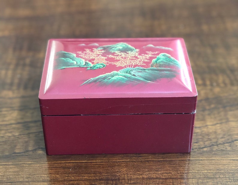 May include: A small, square, red wooden box with a painted scene of green mountains and trees on the lid. The box has a gold trim around the edges.