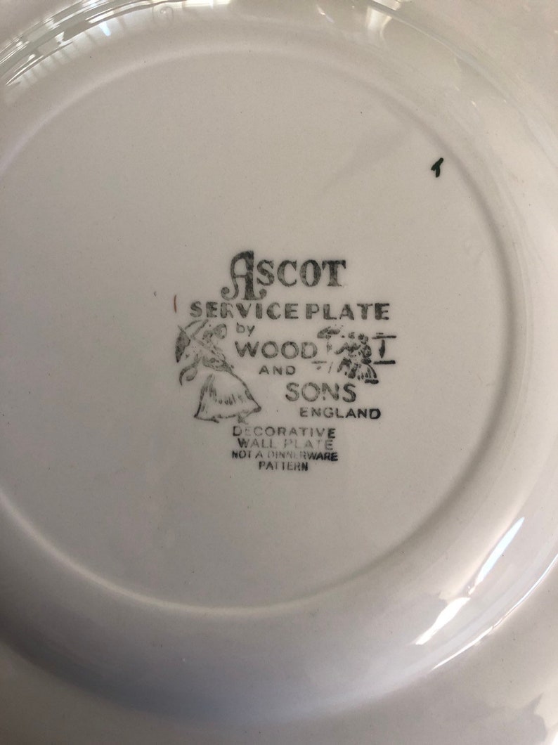 May include: A white ceramic plate with a black and white image of a woman and a man holding umbrellas. The text "ASCOT SERVICE PLATE by WOOD AND SONS ENGLAND DECORATIVE WALL PLATE NOT A DINNERWARE PATTERN" is printed on the plate.