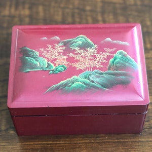 May include: A red wooden box with a hinged lid. The lid is decorated with a painted scene of green mountains, a small pagoda, and trees with gold branches.