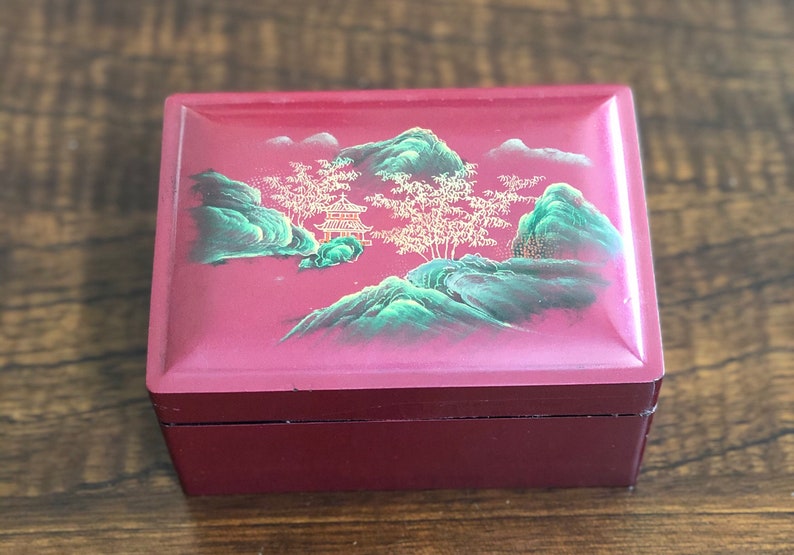 May include: A red wooden box with a hinged lid. The lid is decorated with a painted scene of green mountains, a small pagoda, and trees with gold branches.