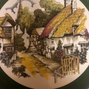 May include: A vintage ceramic plate with a green border features a charming illustration of a quaint English village with thatched-roof cottages, a church steeple, and a winding road.