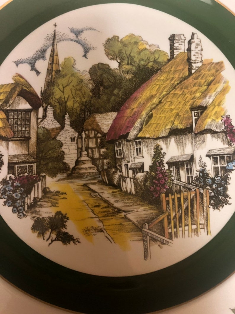 May include: A vintage ceramic plate with a green border features a charming illustration of a quaint English village with thatched-roof cottages, a church steeple, and a winding road.