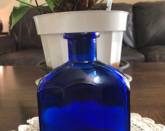 May include: A cobalt blue glass bottle with a rounded, octagonal shape. The bottle is empty and has a smooth, polished surface.