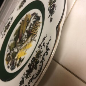 May include: A white ceramic plate with a green border and a gold rim. The plate features a black and white illustration of a cottage with a garden and flowers. The plate is decorated with a black and white floral pattern.