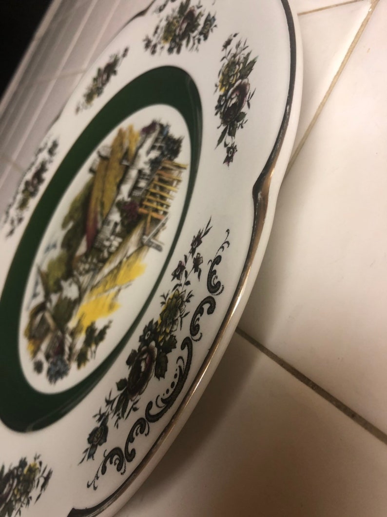 May include: A white ceramic plate with a green border and a gold rim. The plate features a black and white illustration of a cottage with a garden and flowers. The plate is decorated with a black and white floral pattern.