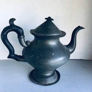 May include: A vintage pewter teapot with a black handle and a lid with a decorative knob. The teapot has a rounded body and a flared spout.