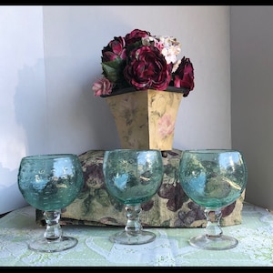 Puede incluir: Tres copas de vidrio azul verdoso con un diseño texturizado de burbujas sobre un mantel de encaje blanco. Las copas tienen una copa redonda y un tallo corto.