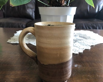 May include: A brown ceramic mug with a light brown glaze and a dark brown base. The mug has a handle and is sitting on a wooden table.