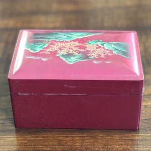 May include: A red wooden box with a hinged lid. The lid is decorated with a painted scene of green mountains and trees in gold.