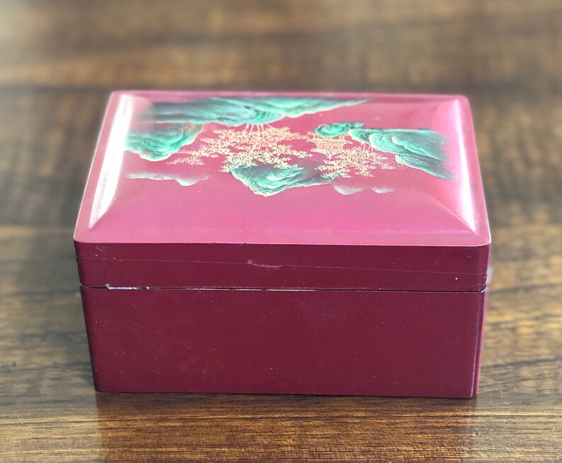 May include: A red wooden box with a hinged lid. The lid is decorated with a painted scene of green mountains and trees in gold.