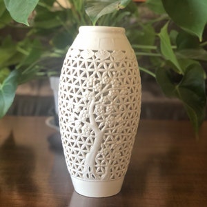 Puede incluir: Jarrón de cerámica blanquecina con un diseño de celosía y un motivo de árbol tallado. El jarrón tiene forma cilíndrica con una base ligeramente ensanchada y un borde decorativo. El diseño del árbol presenta delicadas ramas y flores.