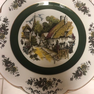 May include: A white ceramic plate with a green border and a floral design. The center of the plate features a black and white illustration of a quaint village with a church steeple.