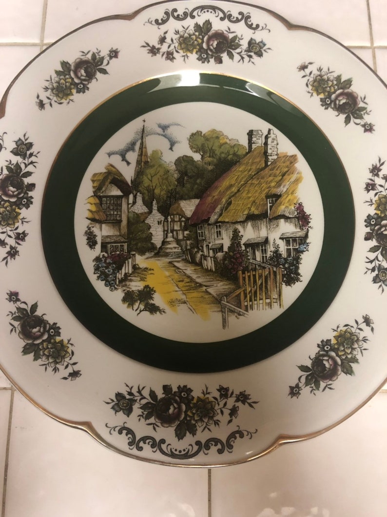 May include: A white ceramic plate with a green border and a floral design. The center of the plate features a black and white illustration of a quaint village with a church steeple.
