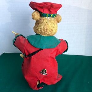 May include: A wind-up toy bear dressed in a red and green outfit with a red hat. The bear is holding a stick in its right paw and has a wind-up key on its back.