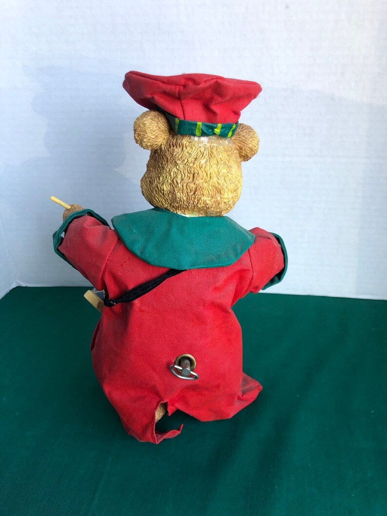 May include: A wind-up toy bear dressed in a red and green outfit with a red hat. The bear is holding a stick in its right paw and has a wind-up key on its back.