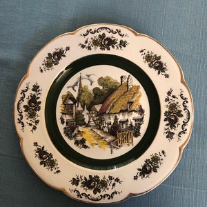 May include: A white ceramic plate with a green border and a floral design. The center of the plate features a vintage illustration of a quaint village with thatched roof cottages and a church steeple.