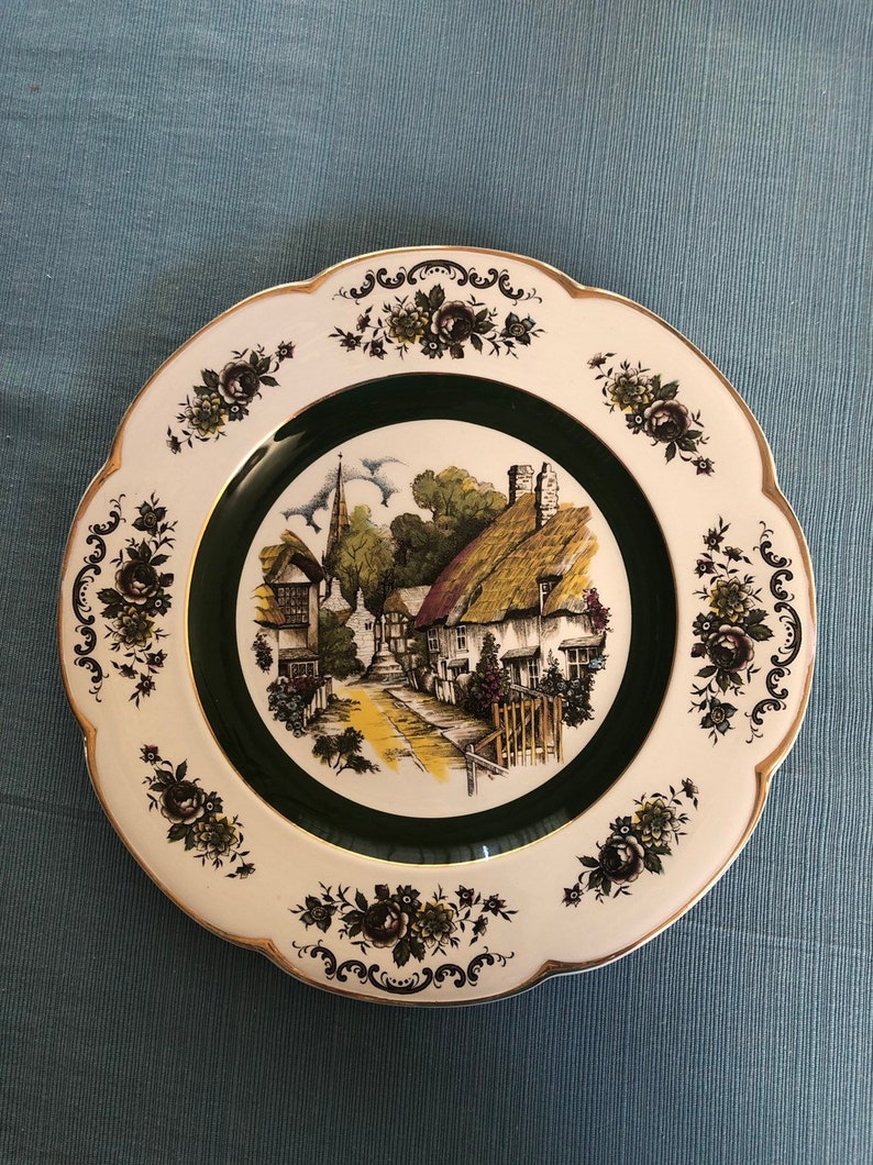 May include: A white ceramic plate with a green border and a floral design. The center of the plate features a vintage illustration of a quaint village with thatched roof cottages and a church steeple.