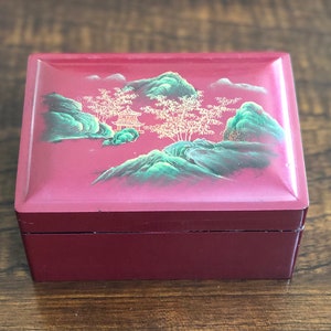May include: A small, red wooden box with a painted scene of a green mountain range and a gold pagoda. The box has a hinged lid and a smooth, lacquered finish.