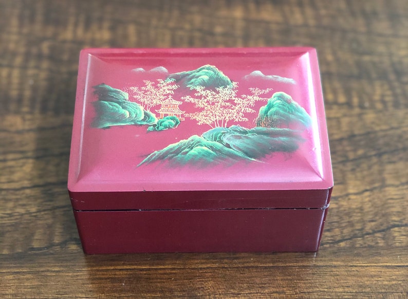 May include: A small, red wooden box with a painted scene of a green mountain range and a gold pagoda. The box has a hinged lid and a smooth, lacquered finish.