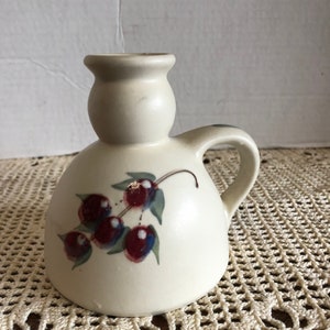 May include: A white ceramic pitcher with a handle and a bulbous body. The pitcher has a painted design of red berries and green leaves.