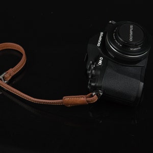 Puede incluir: Una c&aacute;mara Olympus negra con una correa de cuero marr&oacute;n. La c&aacute;mara tiene una tapa de lente con el logotipo de Olympus.