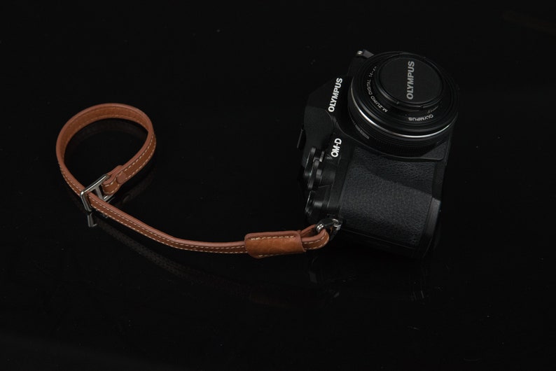 Puede incluir: Una c&aacute;mara Olympus negra con una correa de cuero marr&oacute;n. La c&aacute;mara tiene una tapa de lente con el logotipo de Olympus.