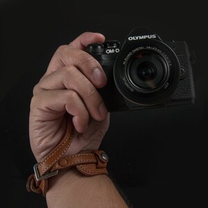 Puede incluir: Una c&aacute;mara Olympus OM-D negra con una correa de mu&ntilde;eca de cuero marr&oacute;n. El objetivo de la c&aacute;mara es un objetivo M.Zuiko Digital 14-42mm 1:3.5-5.6.