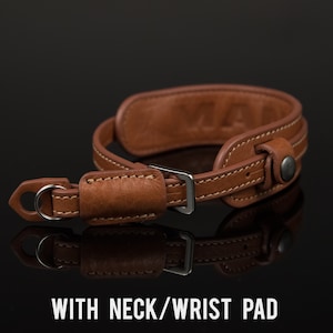 May include: A brown leather camera strap with a metal buckle and a padded neck/wrist strap. The strap has a stitched design and the text "VIA" is embossed on the strap.
