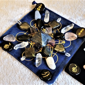 Può includere: Una pochette in velluto blu con un motivo geometrico dorato contiene una collezione di cristalli e pietre, tra cui una piramide di lapislazuli, punte di quarzo trasparente, ossidiana nera e quarzo rosa. La pochette include anche pietre circolari nere con simboli dorati.