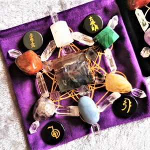 May include: A purple velvet pouch with a variety of crystals and stones arranged in a geometric pattern. The pouch includes clear quartz, amethyst, fluorite, and a blue agate. There are also black stones with gold lettering that say "本" and "卍".
