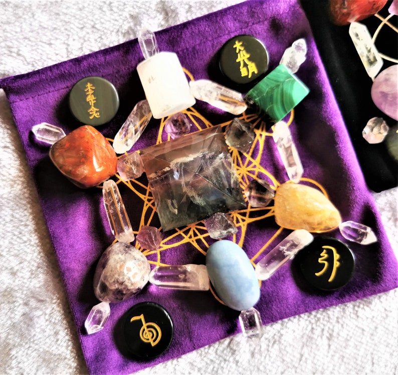 May include: A purple velvet pouch with a variety of crystals and stones arranged in a geometric pattern. The pouch includes clear quartz, amethyst, fluorite, and a blue agate. There are also black stones with gold lettering that say "本" and "卍".