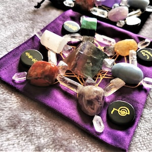 May include: A purple velvet pouch with a variety of crystals and stones arranged in a geometric pattern. The pouch includes clear quartz, amethyst, fluorite, red jasper, citrine, and a blue stone. There are also black circular stones with gold symbols.