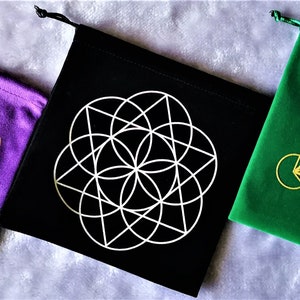May include: Three velvet drawstring pouches with different colored backgrounds: purple, black, and green. Each pouch features a gold geometric design of a flower of life pattern.