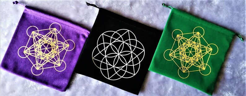 May include: Three velvet drawstring pouches with different colored backgrounds: purple, black, and green. Each pouch features a gold geometric design of a flower of life pattern.